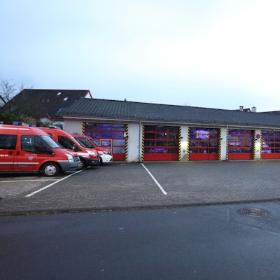 Eine Feuerwache mit roten Rolltoren, links stehen drei Feuerwehrfahrzeuge im Freien.