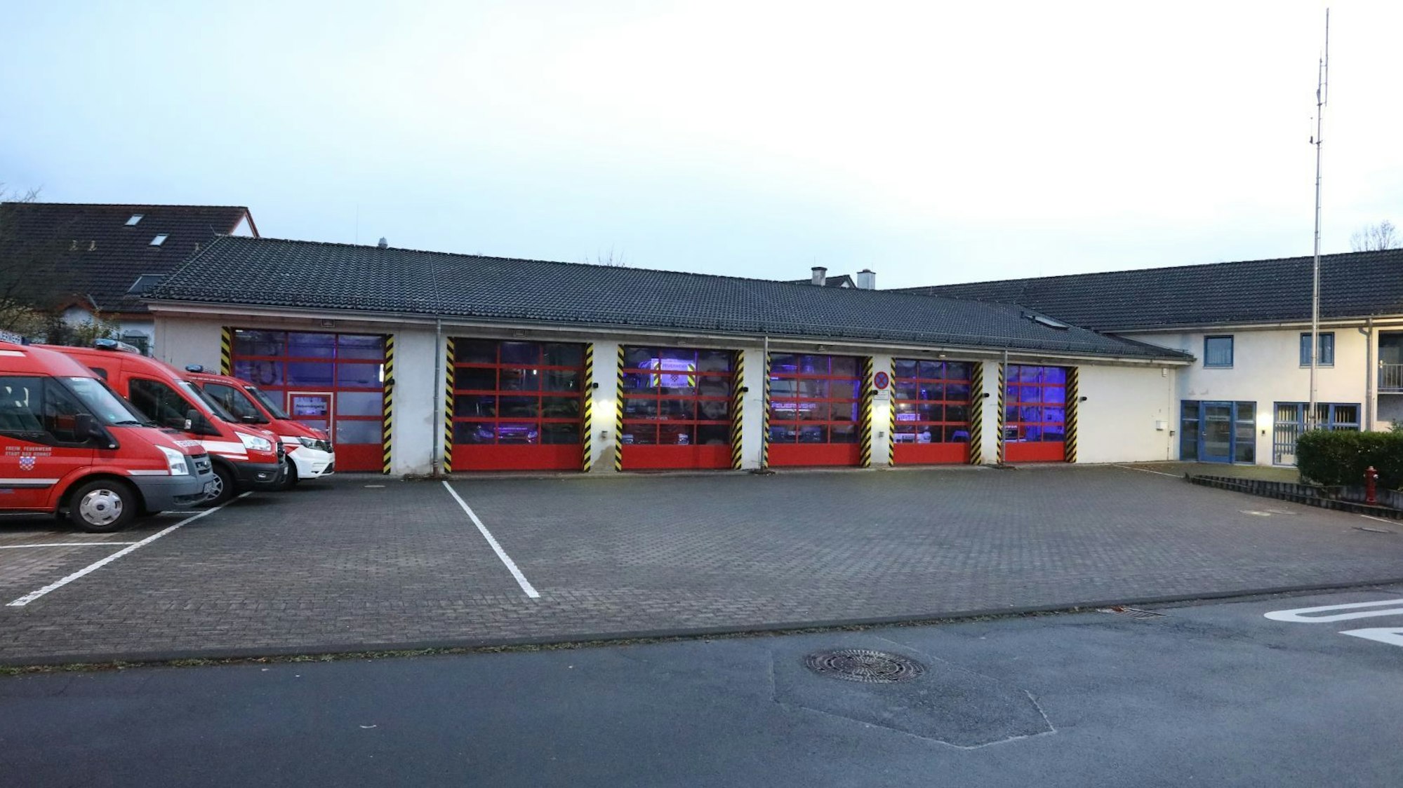 Eine Feuerwache mit roten Rolltoren, links stehen drei Feuerwehrfahrzeuge im Freien.