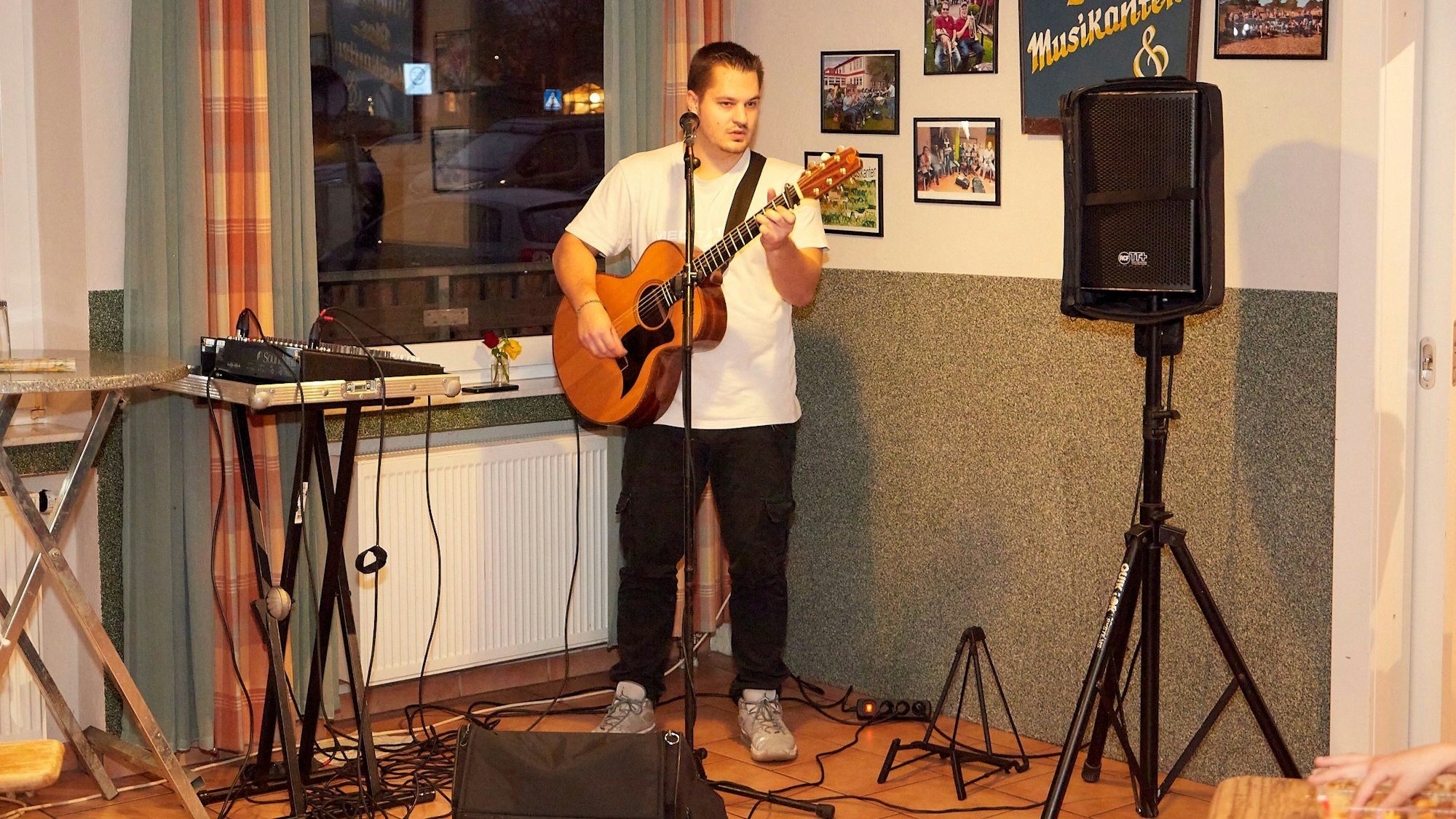 Ein junger Mann in weißem T-Shirt und schwarzer Jeans steht mit einer Gitarre an einem Mikrofon.