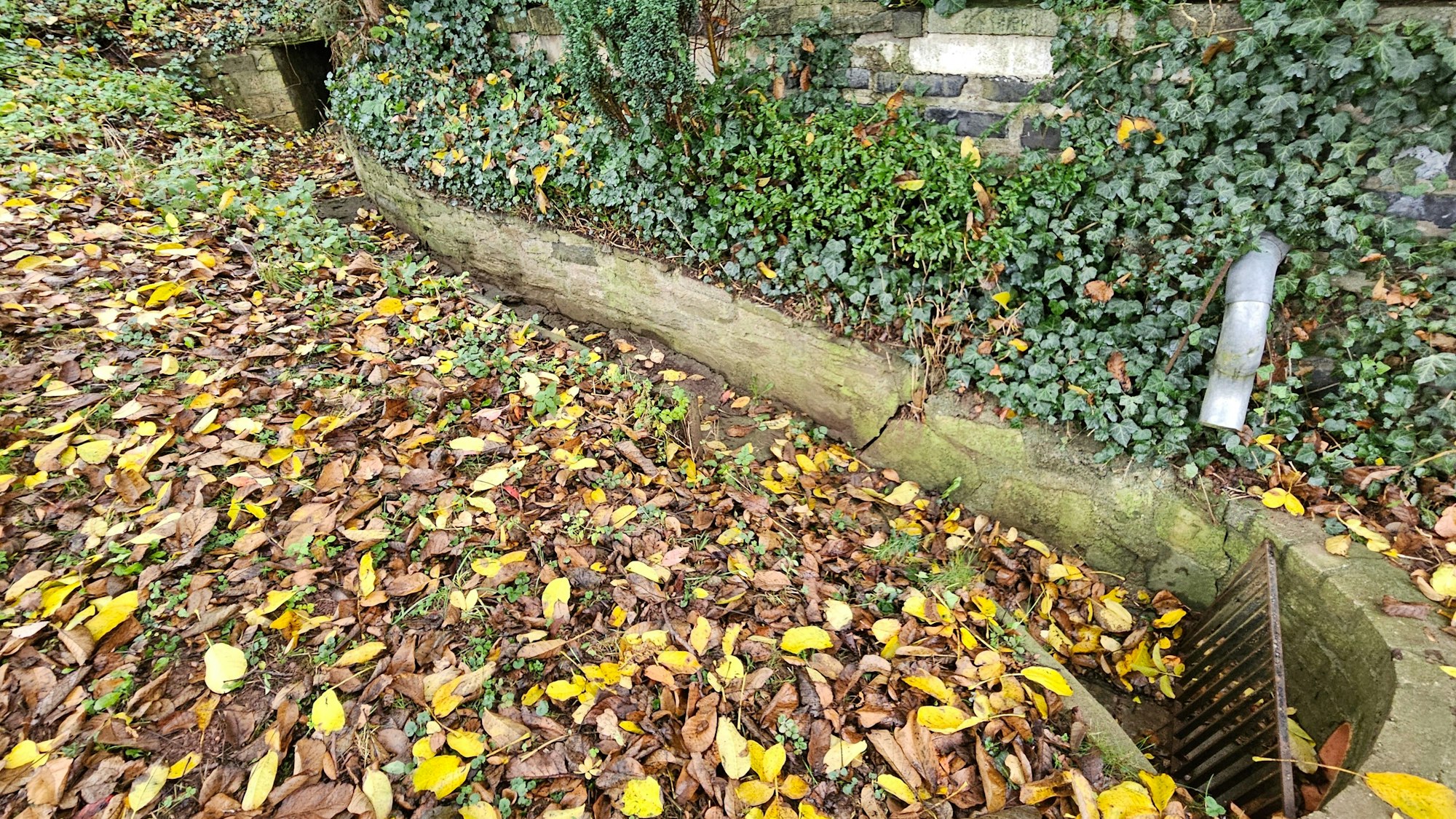 Das Foto zeigt den Durchlass durch den Bahndamm und einen offenen, mit Steinen angelegten Graben. Durch ihn läuft das Wasser in ein Rohr, das durch ein Gitter geschützt ist.