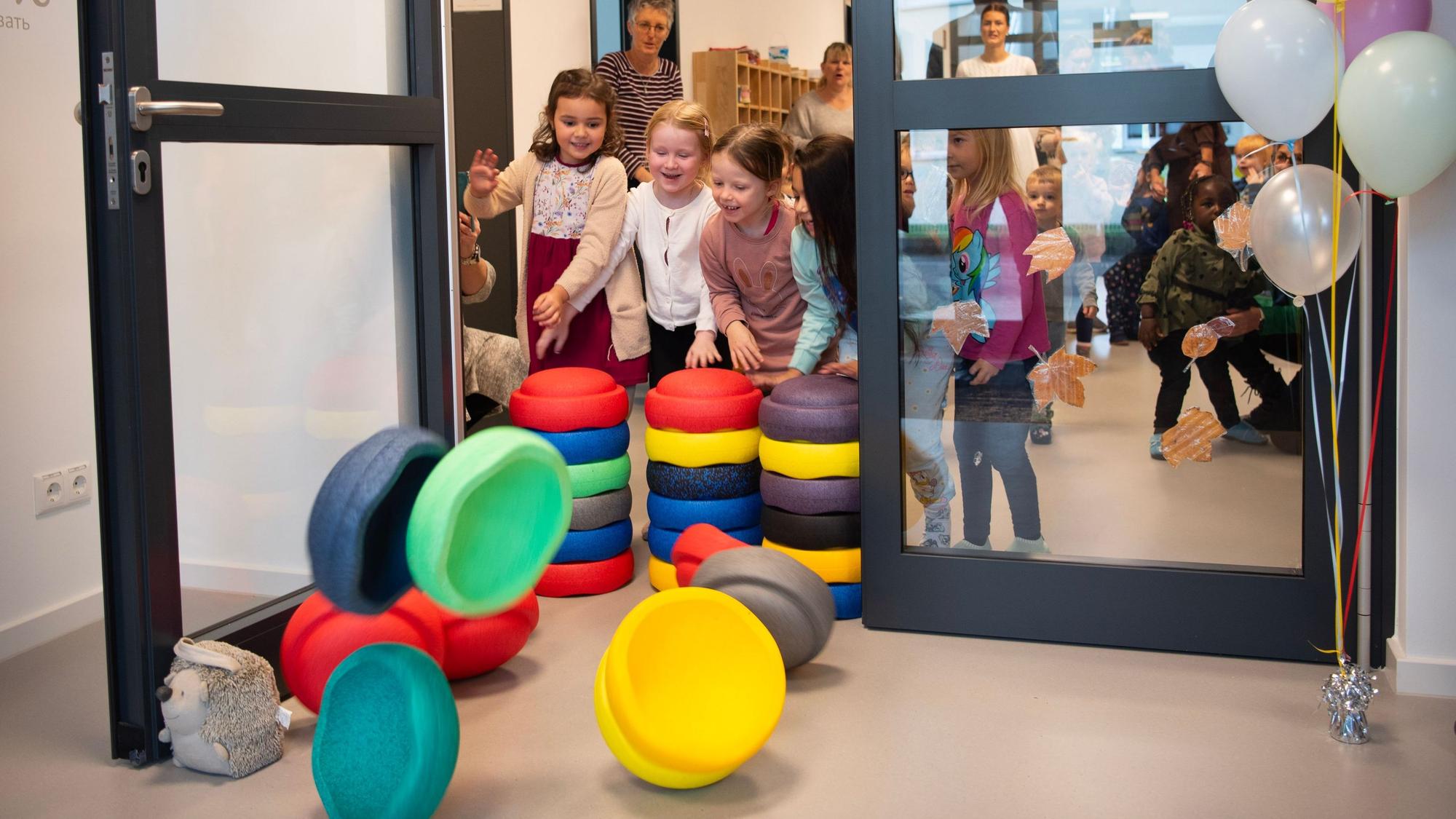 Kinder schmeißen bunte Stapelsteine, die eine Tür versperren, um. Neben der Tür steht ein Strauß bunter Luftballons.