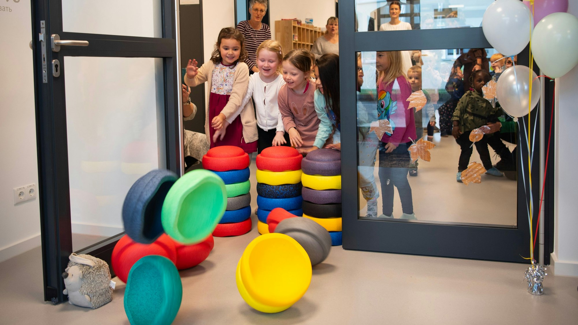 Kinder schmeißen bunte Stapelsteine, die eine Tür versperren, um. Neben der Tür steht ein Strauß bunter Luftballons.