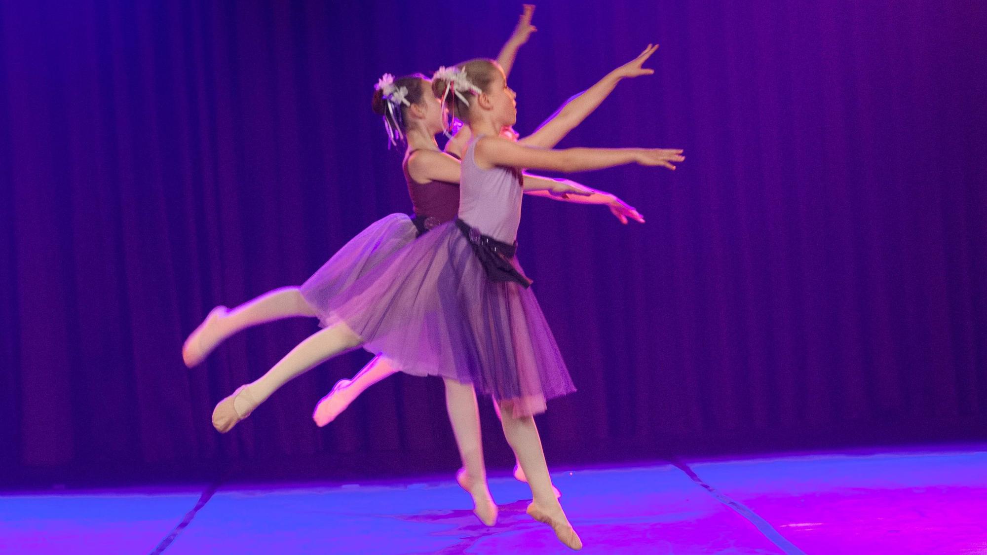 Zwei junge Ballett-Tänzerinnen strecken in lila Kostümen ihre Extremitäten formvollendet von sich.