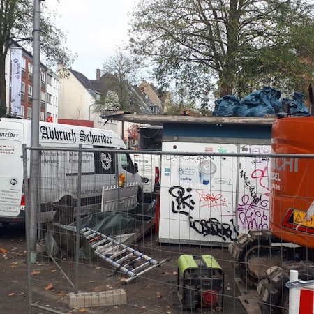 Hinter einem Bauzaun steht schweres Gerät und ein kleines flaches Gebäude.