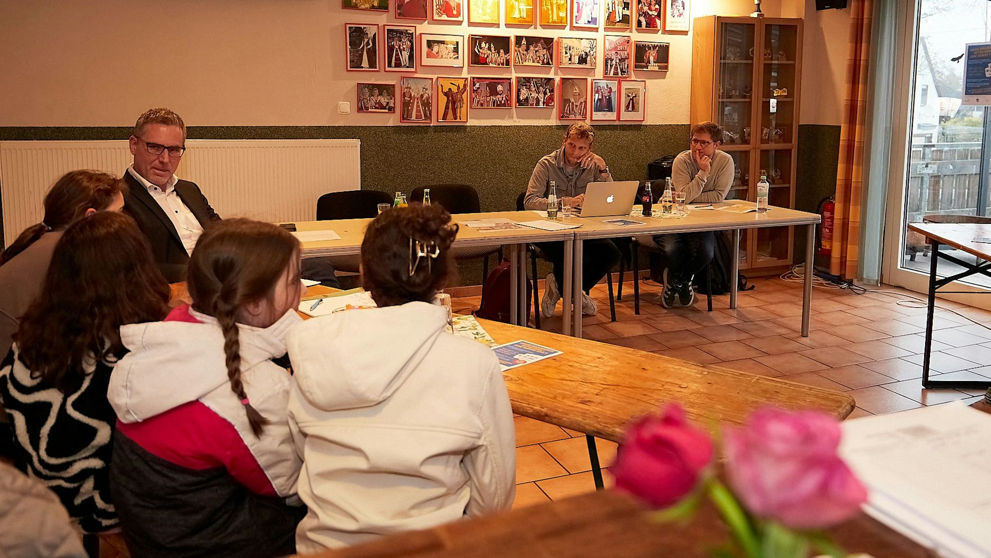 Mehrere junge Menschen sitzen an einem Biertisch (links im Bild), mit ihnen sprechen mehrere Erwachsene, die ebenfalls an Tischen sitzen. Auf den Tischen stehen Wasser- und Cola-Flaschen, an den Wänden hängen Fotos.
