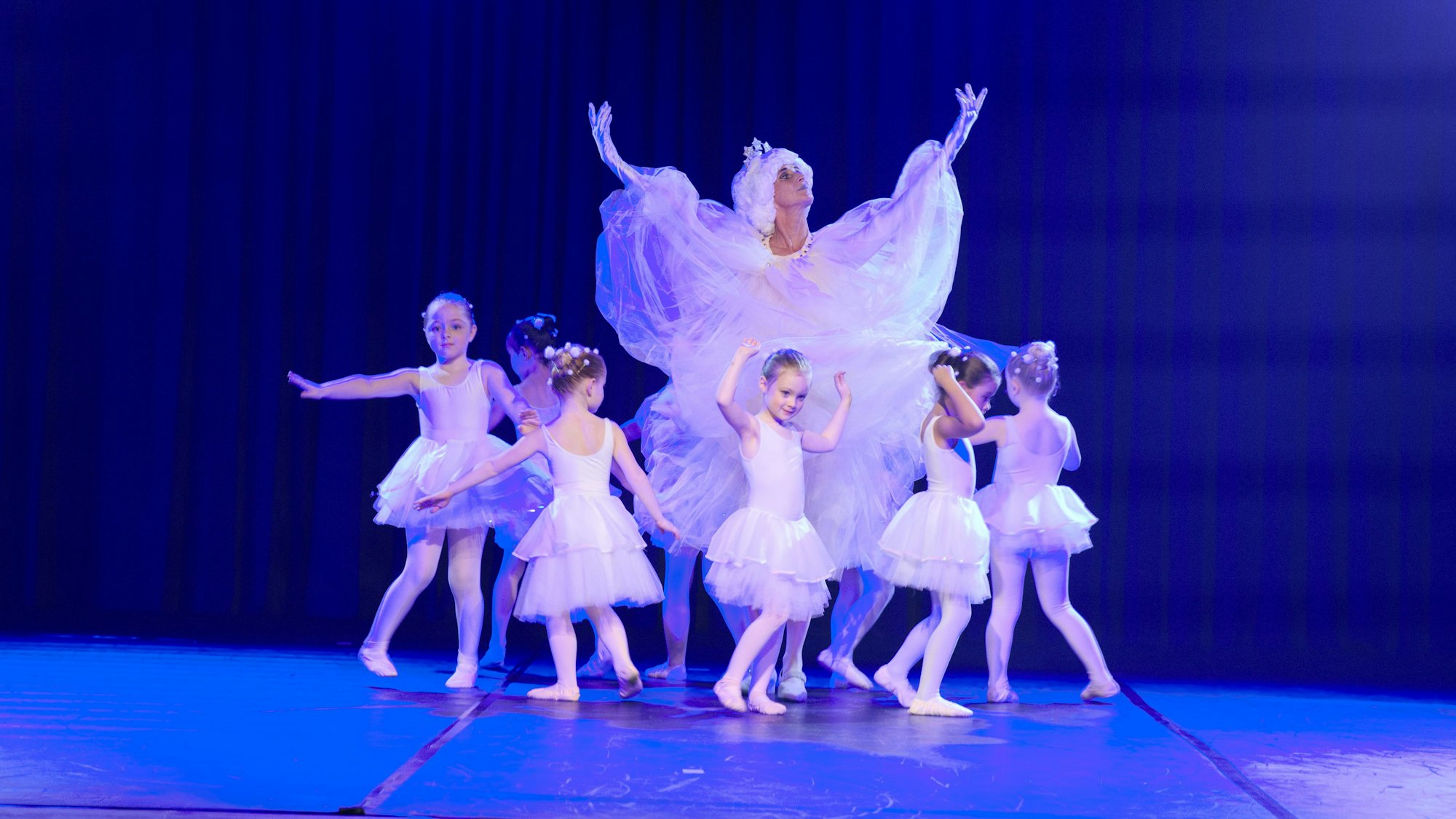 Junge Ballett-Tänzerinnen umkreisen in weißen Tütüs eine Frau in weißem Kostüm mit Krone.