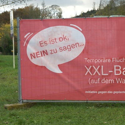 Ein großes, rotes Plakat steht auf einer Wiese. Darauf ist in einer Sprechblase zu lesen: "Es ist okay, NEIN zu sagen...". Und: "Temporäre Flüchtlingsunterkunft JA - XXL-Bau NEIN". Initiative gegen das geplante Bauvorhaben in Odenthal-Höffe.