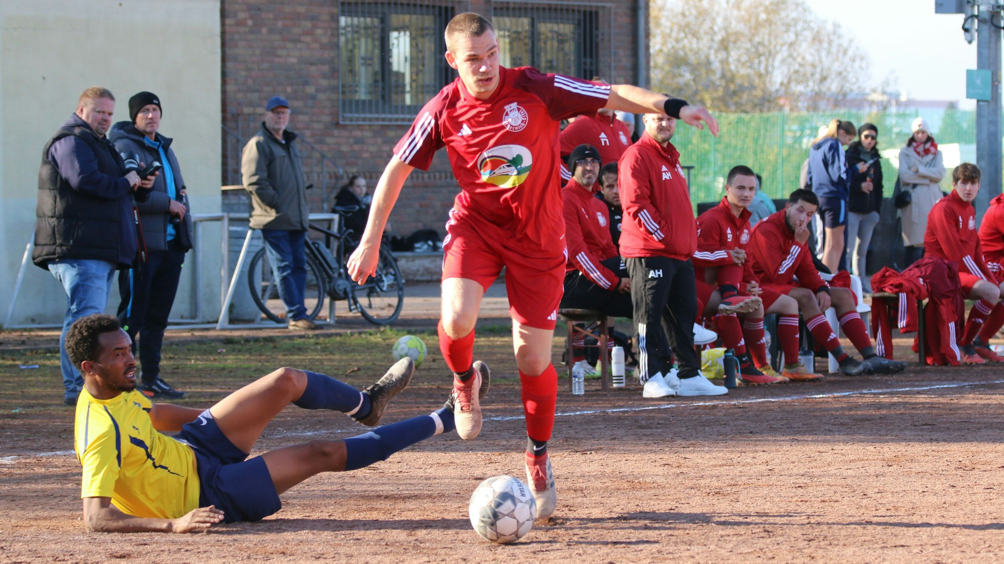 Ein Fußballer der TuS Mechernich legt einen Ball am auf dem Boden liegenden Spieler aus Kommern vorbei.