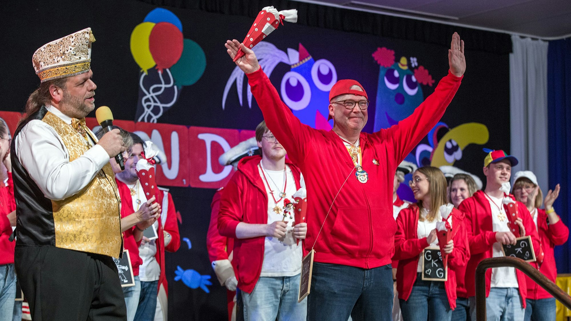 Auf einer Bühne in Worringen stehen einige rot-weiß-gekleidete Menschen. Sie feiern Sessionseröffnung. Der Moderator trägt goldene Weste und Narrenkappe.