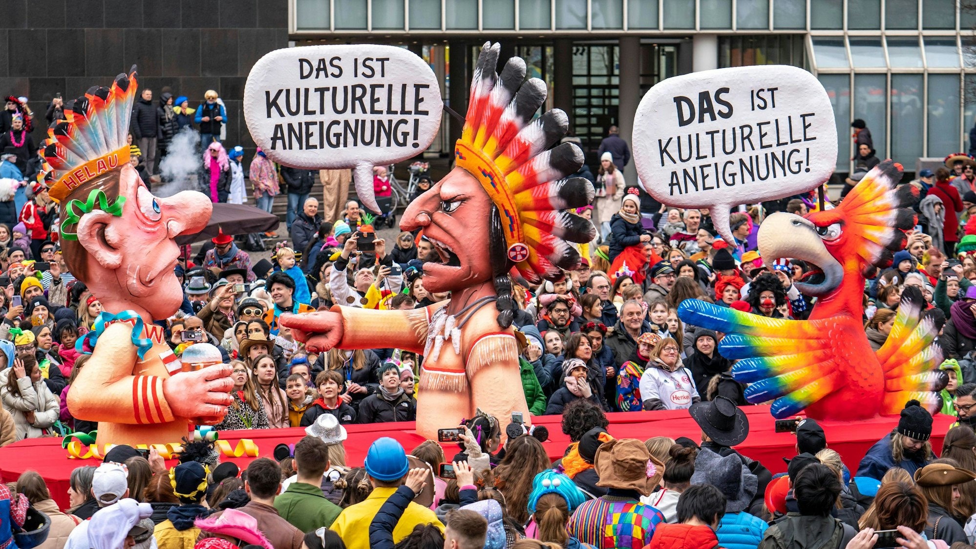 Ein Mottowagen des Düsseldorfer Karnevalszugs 2023 verbildlicht das Thema „kulturelle Aneignung“ im Karneval, indem eine indigene Figur mit Federkopfschmuck einen als Indianer verkleideten Jecken der „kulturellen Aneignung“ beschuldigt, er aber gleichzeitig von einem Vogel mit buntem Federkleid desselben Vergehens beschuldigt wird.