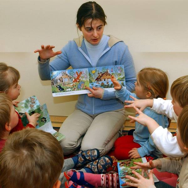 Eine Erzieherin liest mit Kindern in einer Kita ein Bilderbuch, sie sitzt vor den Kindern und zeigt ihnen die Bilder, dazu gestikulieren alle mit den Händen.
