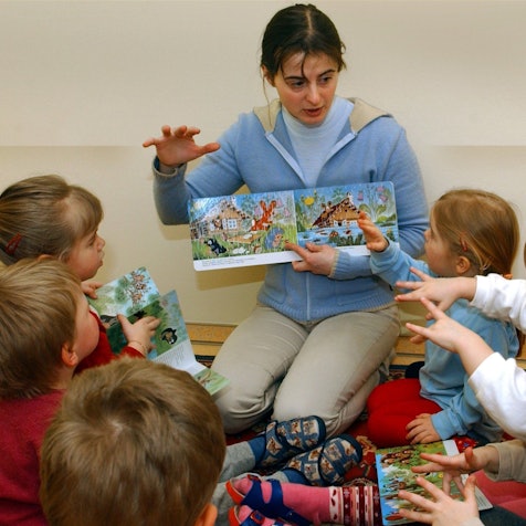 Eine Erzieherin liest mit Kindern in einer Kita ein Bilderbuch, sie sitzt vor den Kindern und zeigt ihnen die Bilder, dazu gestikulieren alle mit den Händen.