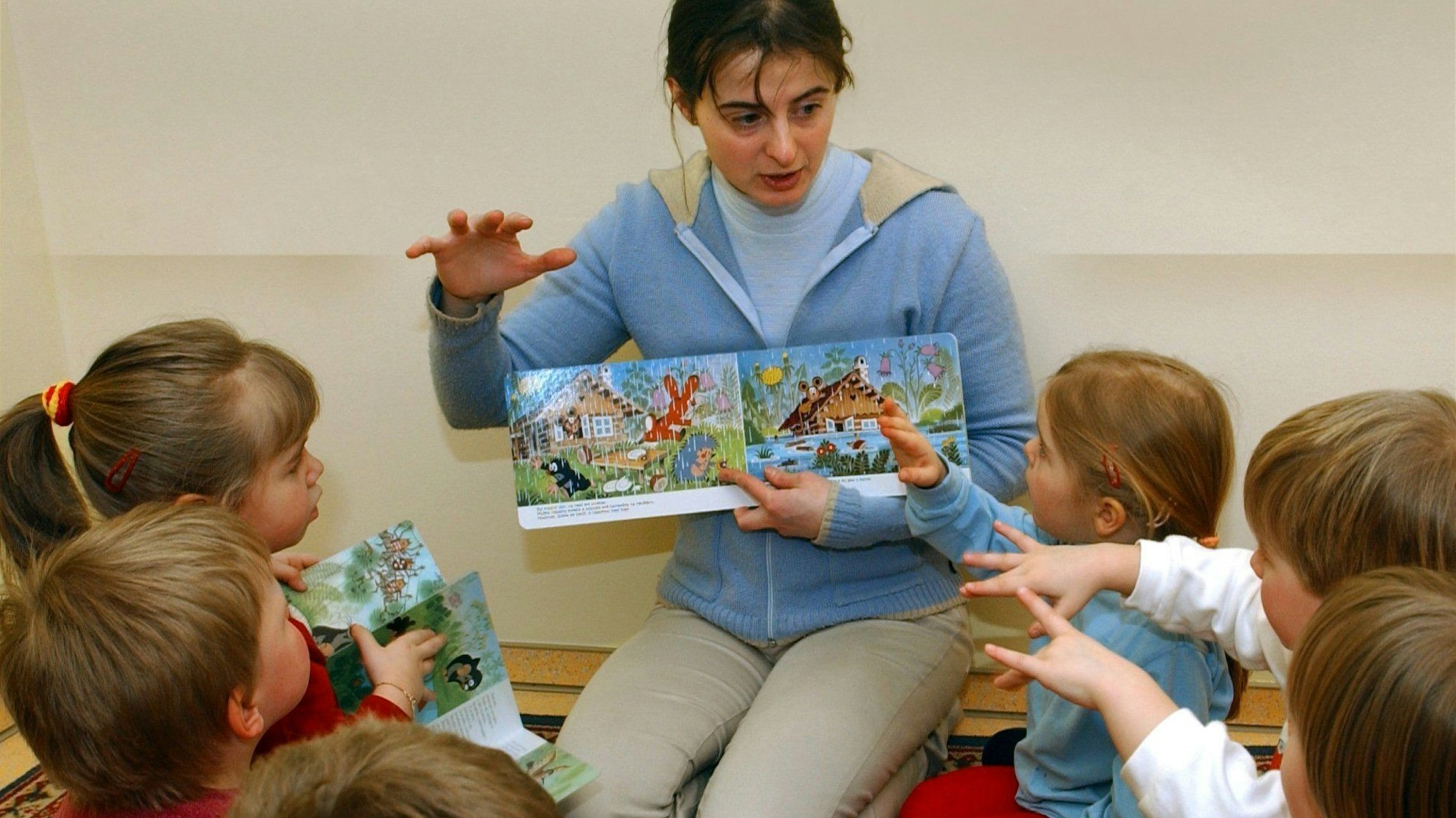 Eine Erzieherin liest mit Kindern in einer Kita ein Bilderbuch, sie sitzt vor den Kindern und zeigt ihnen die Bilder, dazu gestikulieren alle mit den Händen.