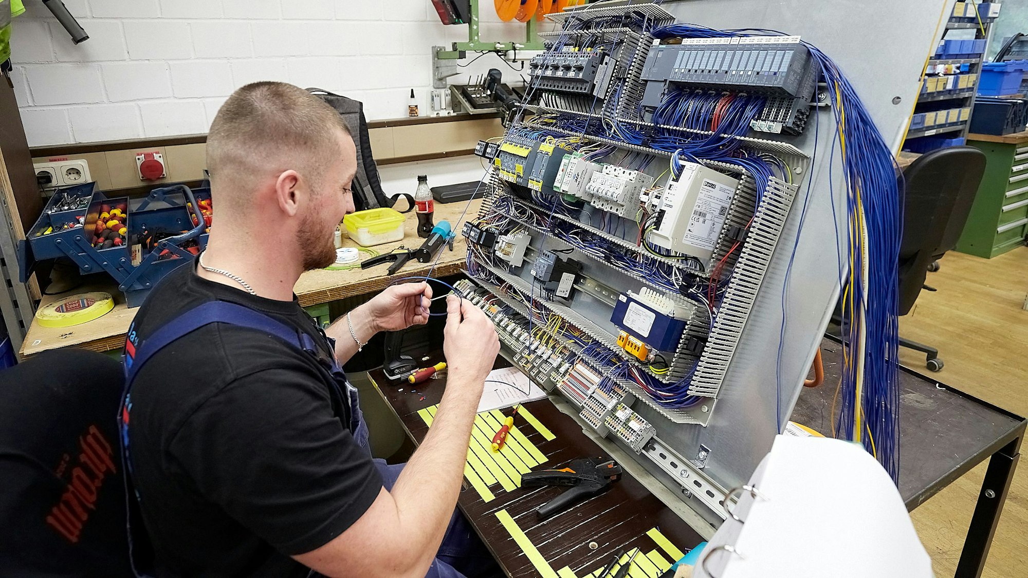 Ein Mann in schwarzem T-Shirt und blauer Latzhose sitzt vor einer grauen Tafel mit vielen Schaltern und Kabeln.