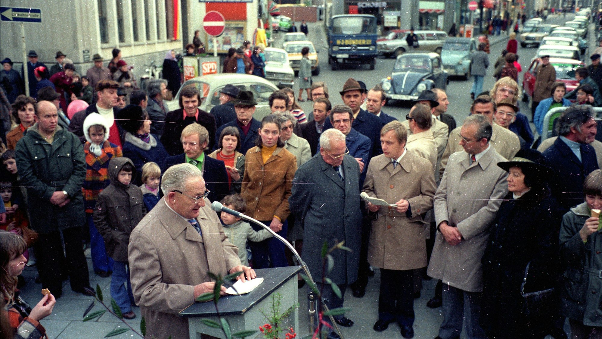 Schlösser spricht an einem Pult in ein Mikrofon, umringt von Zuhörerinnen und Zuhörern.