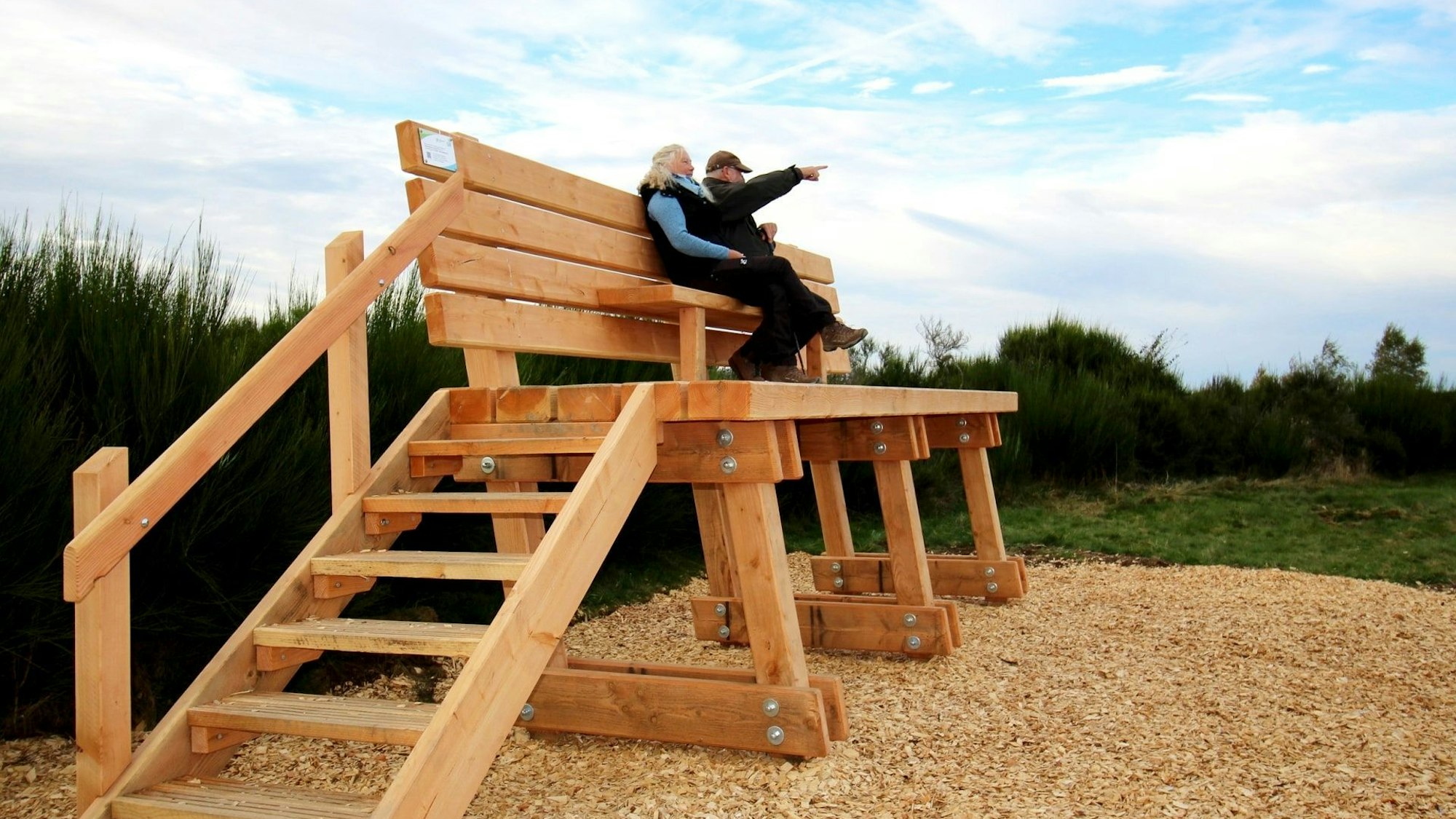 Auf der überdimensionalen Bank sitzen eine Frau und ein Mann. Er streckt seinen rechten Arm aus, um der Frau etwas zu zeigen.