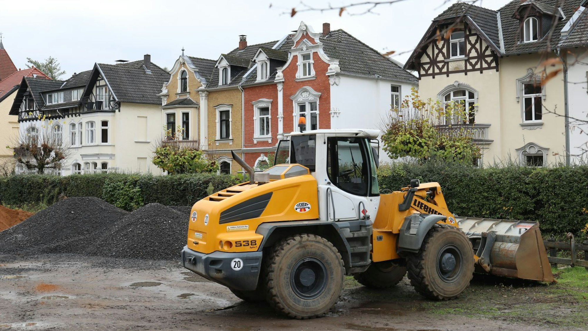 Ein Bagger auf einer matschigen Fläche, im Hintergrund stehen Jugendstilhäuser.