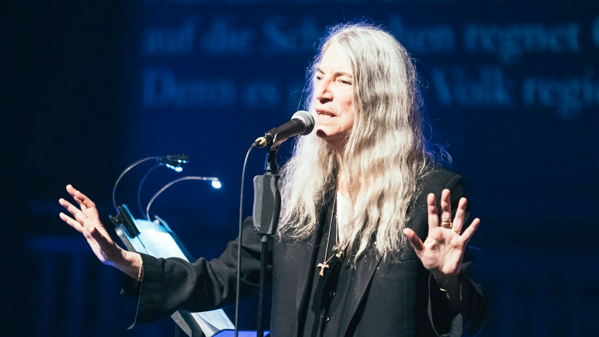 Zu sehen ist die Sängerin Patti Smith auf einer Bühne, vor einem Mikrofon.