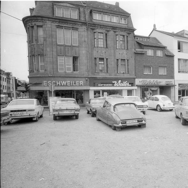 Auf einem Platz stehen Autos, im Hintergrund die Neustraße mit Geschäftslokalen.