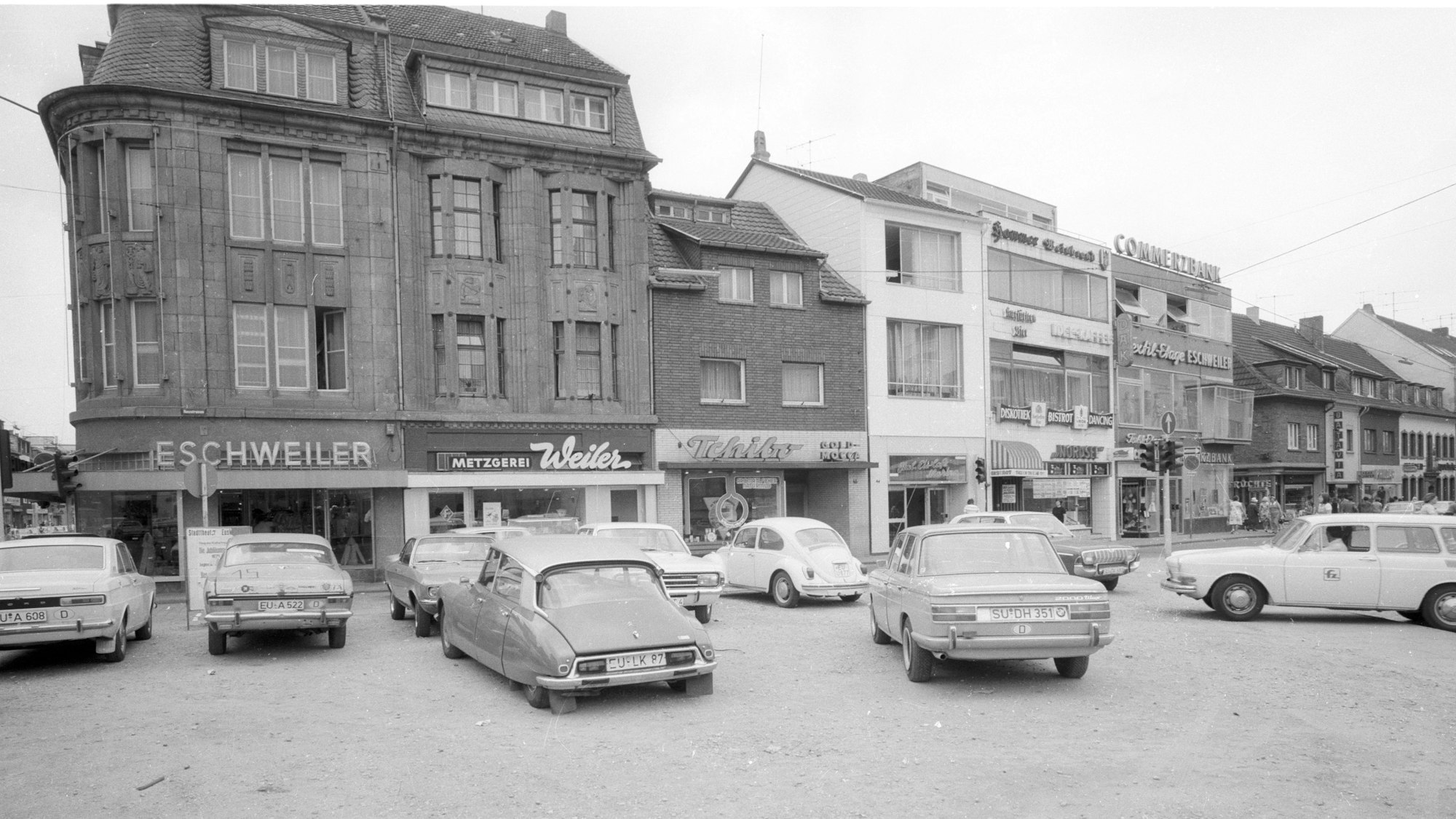 Auf einem Platz stehen Autos, im Hintergrund die Neustraße mit Geschäftslokalen.