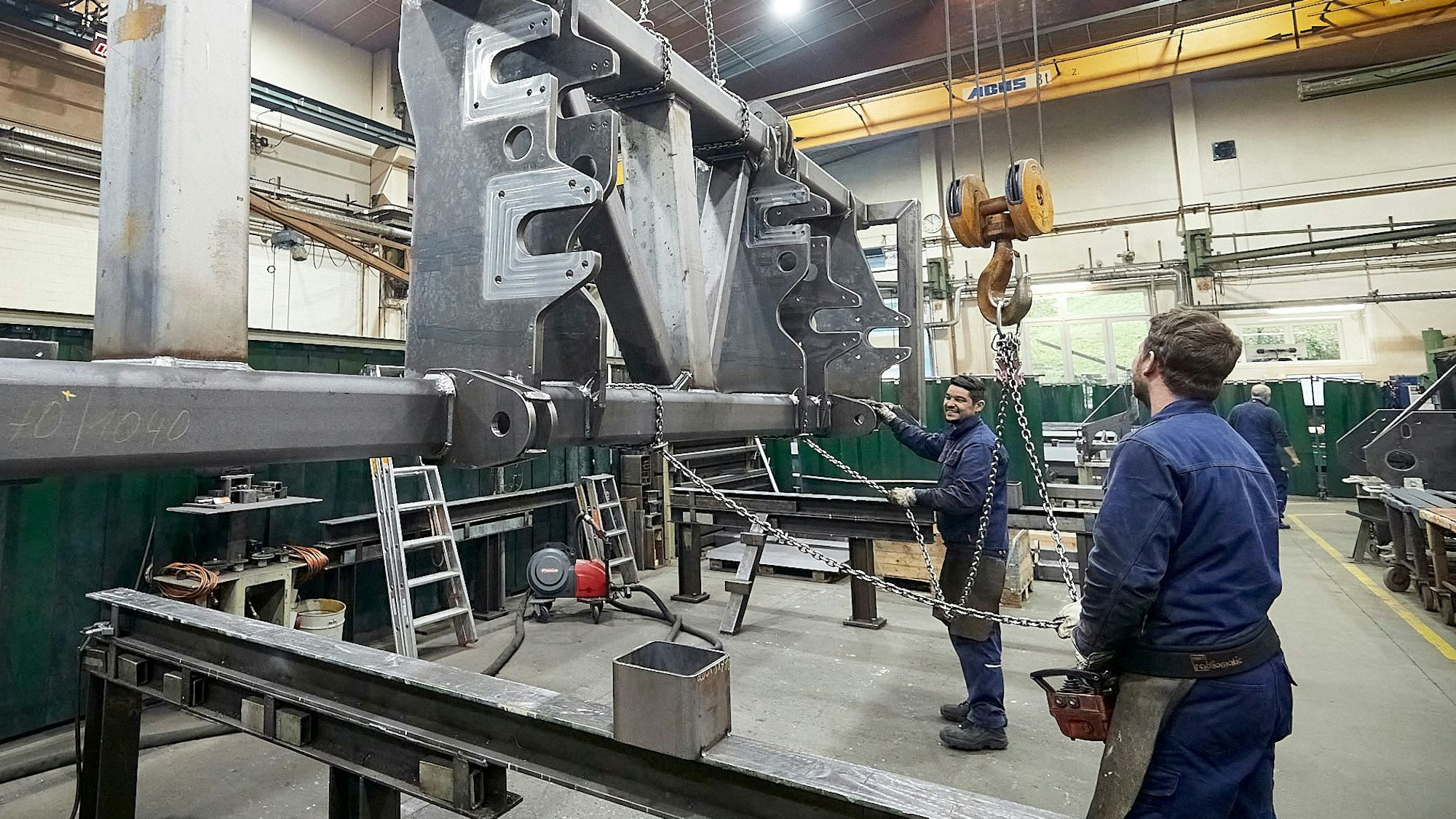 Zwei Männer in blauer Arbeitskleidung stehen in einer großen Halle und bedienen einen Kran, an dem ein großes Stahlteil hängt.