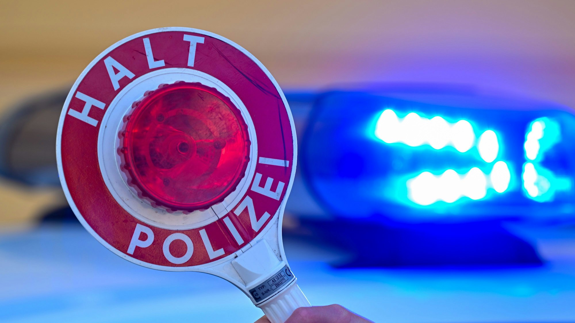 Das Symbolbild zeigt eine Kelle der Polizei mit der Aufschrift „Halt Polizei“ vor dem Blaulicht eines Polizeifahrzeuges.