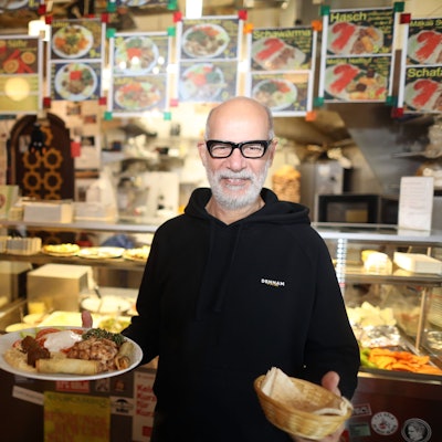Fuad Taih steht vor der Theke des Habibis. In der einen Hand hält er einen Teller mit einem Gericht, in der anderen einen Brotkorb.