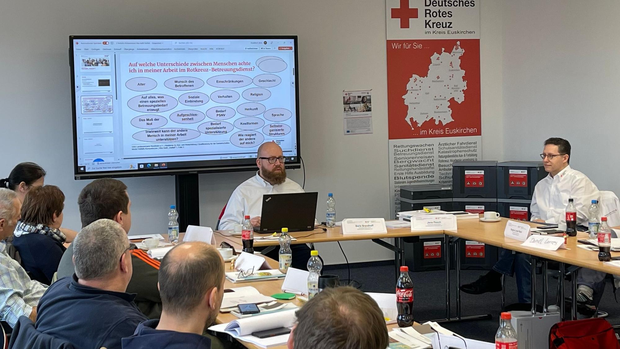 Eine Schulung. Links sitzen die Teilnehmenden an Tischen, vorne bedient ein Mann einen Laptop. Auf dem Smartboard hinter ihm sind Schulungsinhalte zu lesen. Rechts sitzt ein weiterer männlicher Ausbilder.
