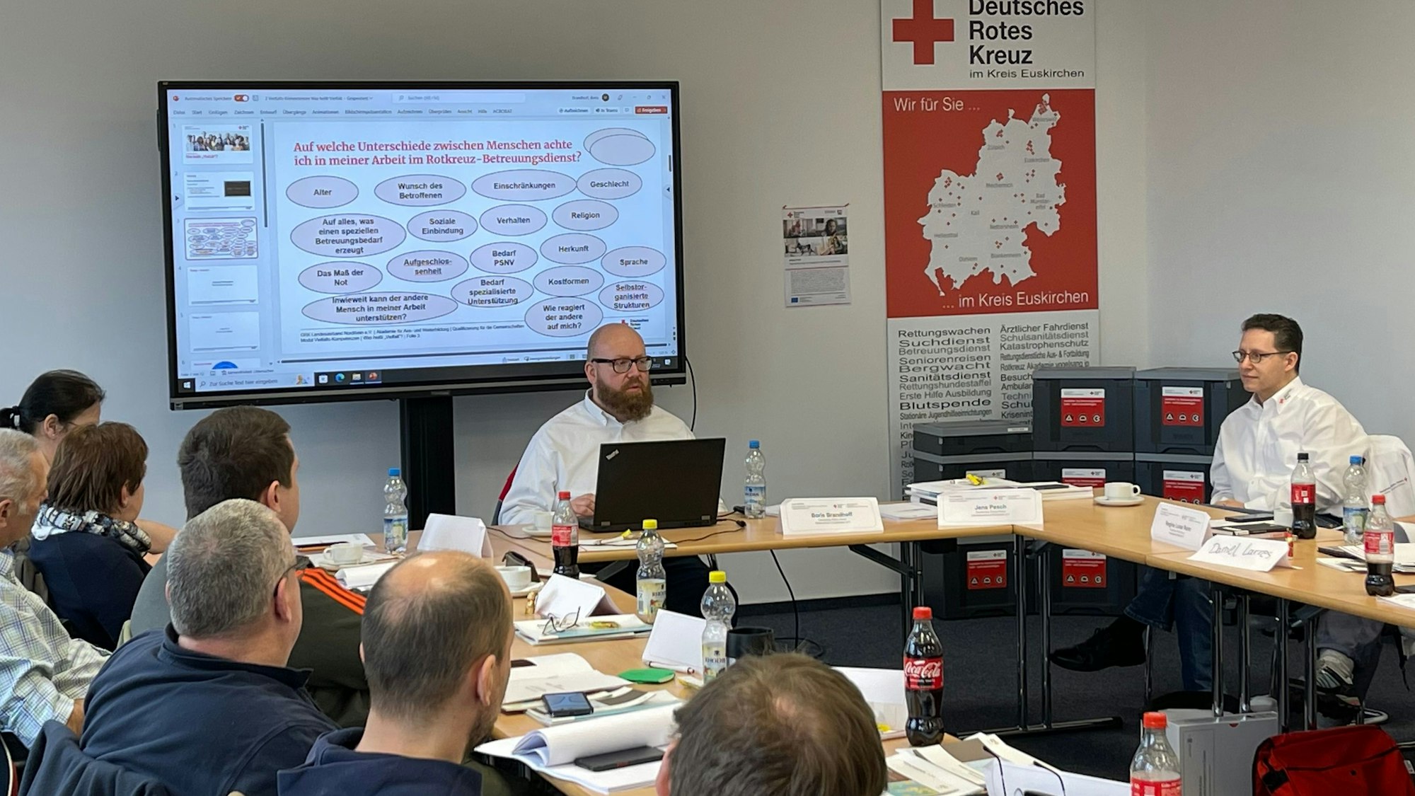 Eine Schulung. Links sitzen die Teilnehmenden an Tischen, vorne bedient ein Mann einen Laptop. Auf dem Smartboard hinter ihm sind Schulungsinhalte zu lesen. Rechts sitzt ein weiterer männlicher Ausbilder.