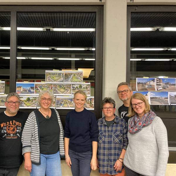 Sieben Menschen stehen fröhlich vor Glasscheiben, an denen Bilder hängen.