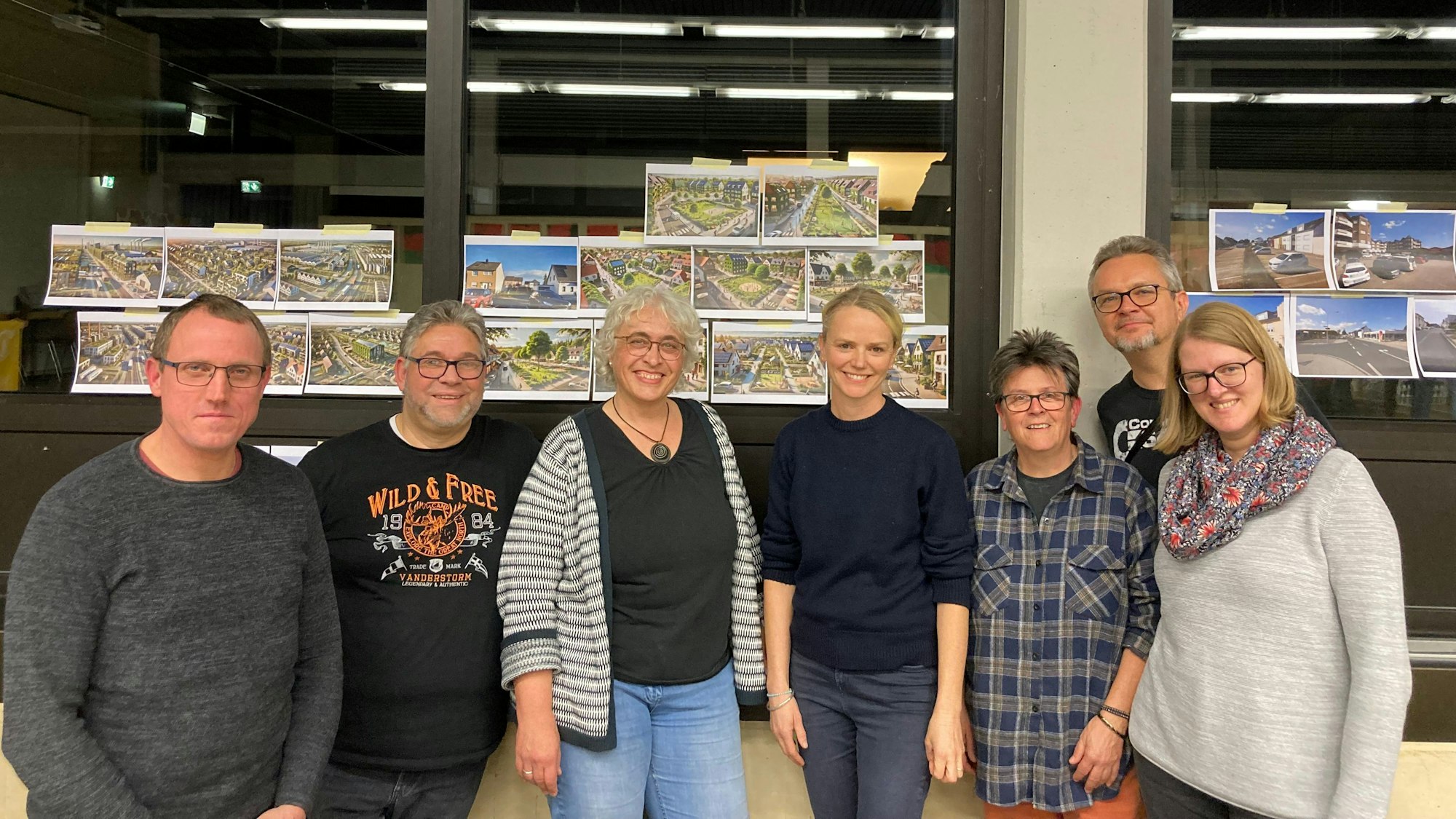 Sieben Menschen stehen fröhlich vor Glasscheiben, an denen Bilder hängen.
