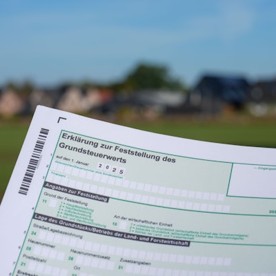 Eine Erklärung zur Feststellung des Grundsteuerwerts für das Jahr 2025 wird vor eine Wohnsiedlung hochgehalten.