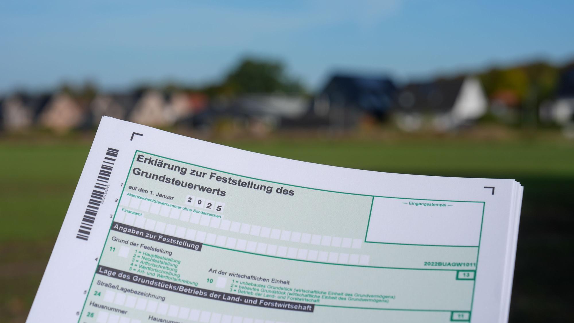 Ein Formular mit der Erklärung zur Feststellung des Grundsteuerwerts für das Jahr 2025 wird vor eine Wohnsiedlung hochgehalten.