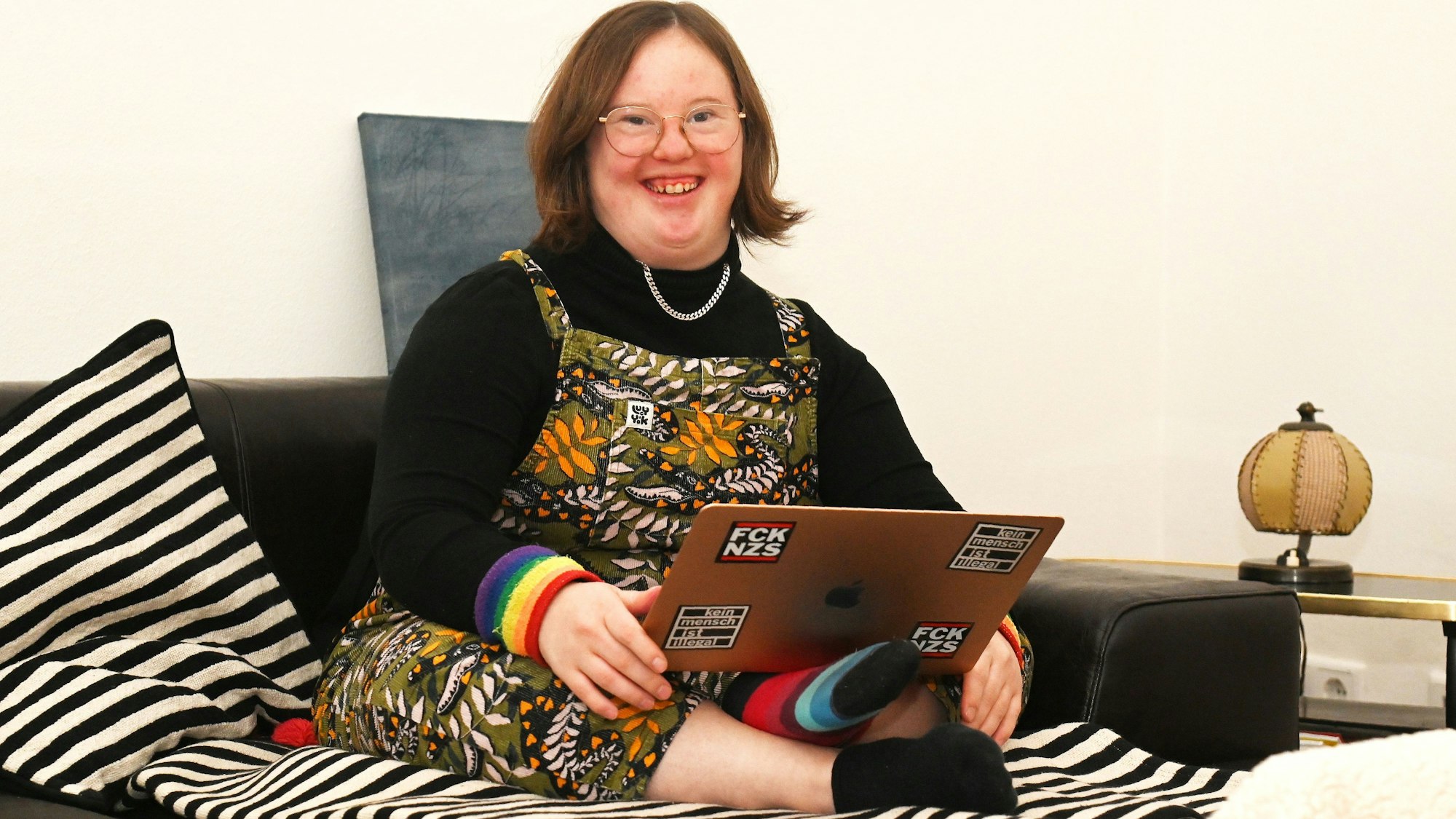 Eine junge Frau sitzt im Schneidersitz auf dem Sofa, auf den Knien hat sie einen Laptop.