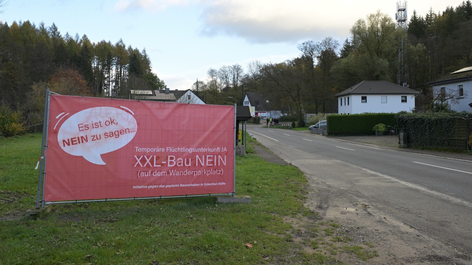 Auf einem roten Plakat am Straßenrand spricht sich eine Bürgerinitiative gegen den Bau eines Flüchtlingsheims in Odenthal-Höffe aus.