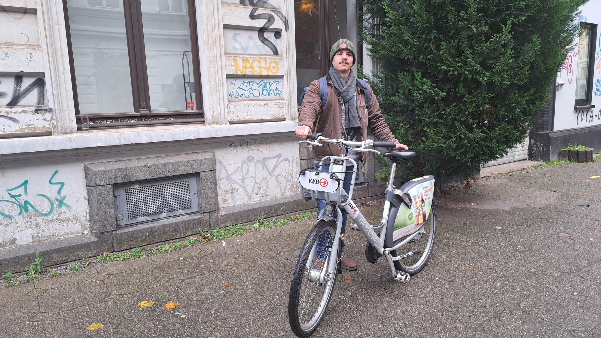 Fabio Botero steht mit einem KVB-Rad auf dem Gehweg.