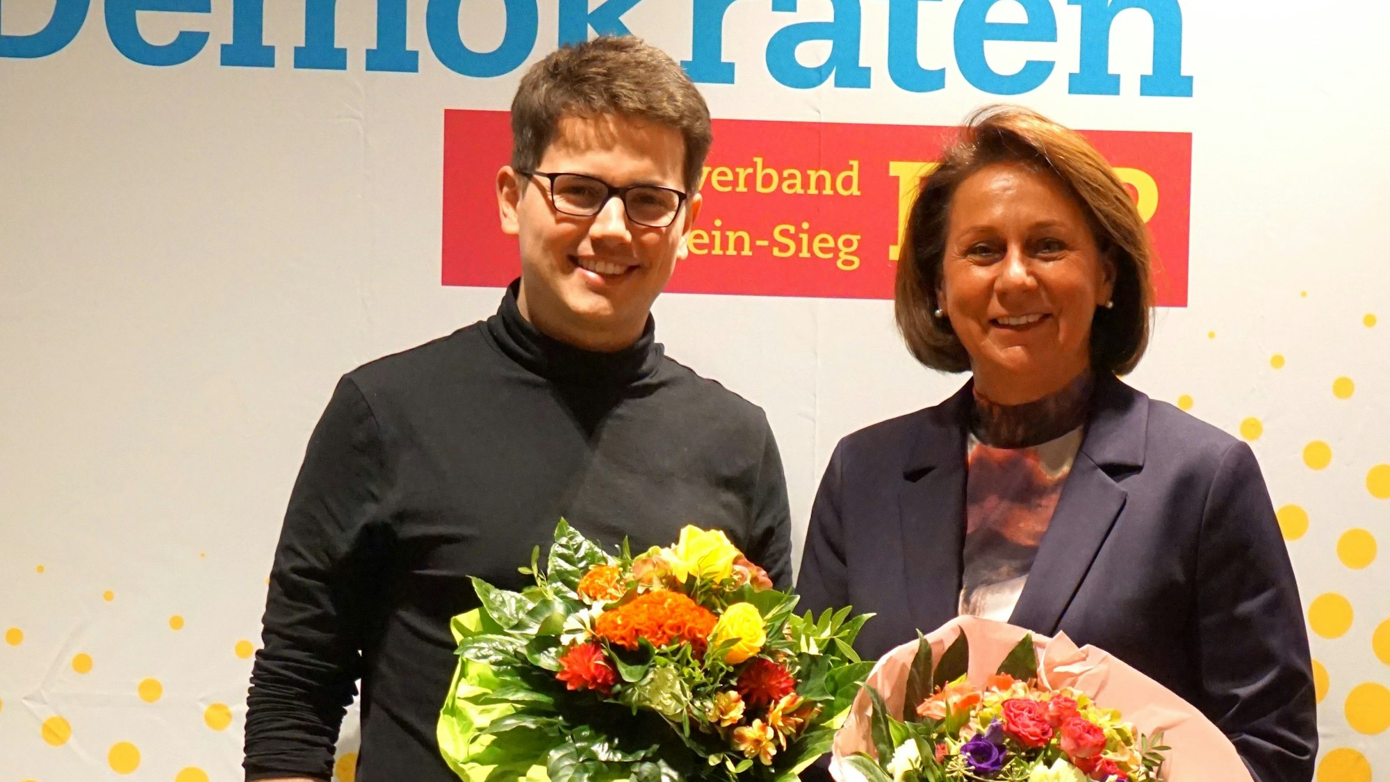 Ein Mann und eine Frau stehen mit Blumensträußen vor einer Wand mit dem Schriftzug der FDP Rhein-Sieg