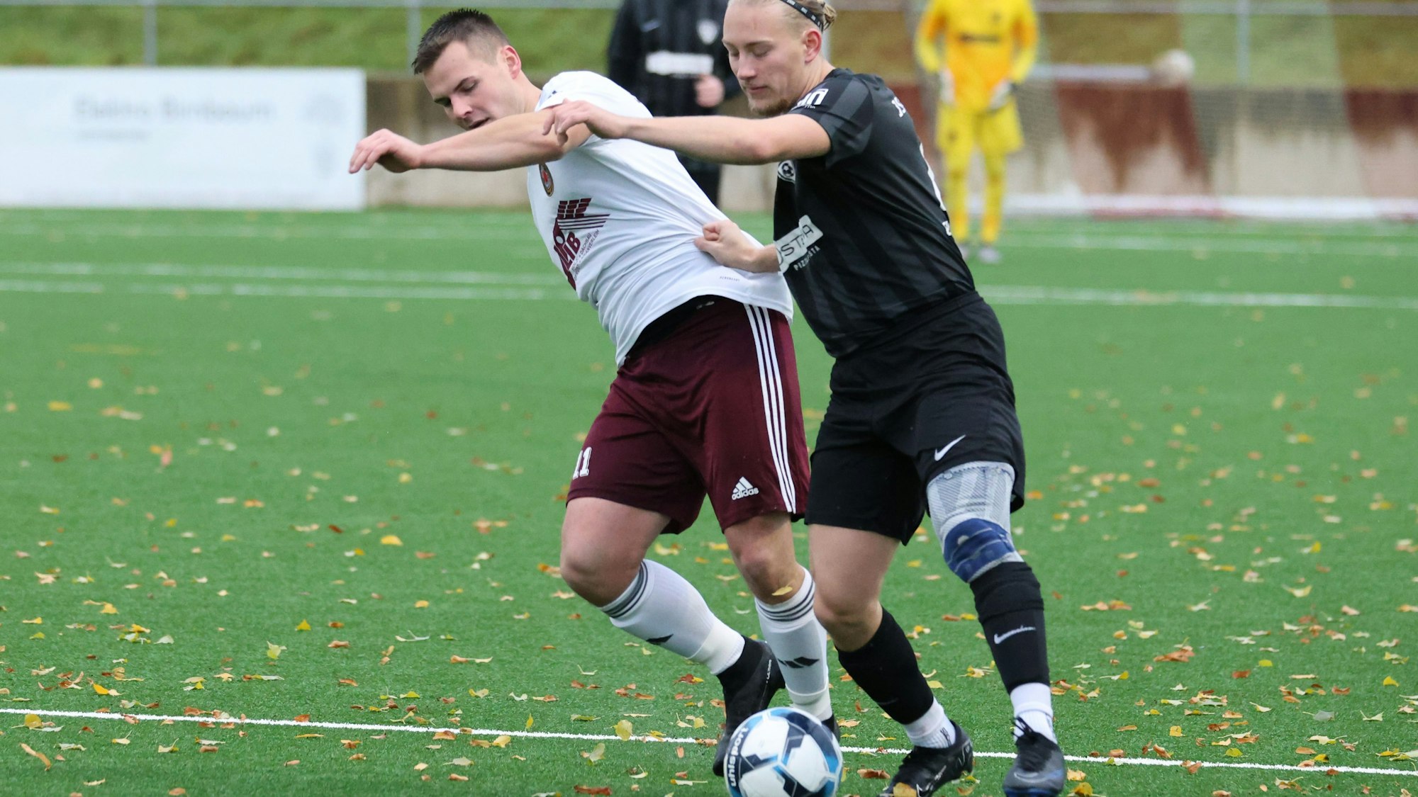 Zweikampf zwischen zwei Fußballern. Auf dem Kunstrasenplatz liegt Laub.