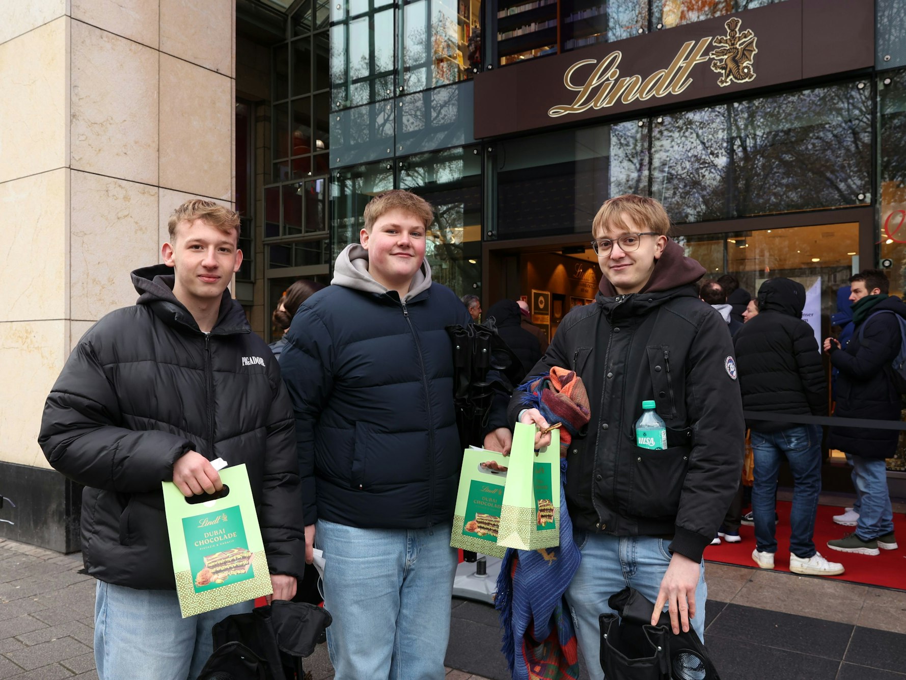 Drei junge Männer stehen mit Papiertüten vor einer Lindt-Filiale.
