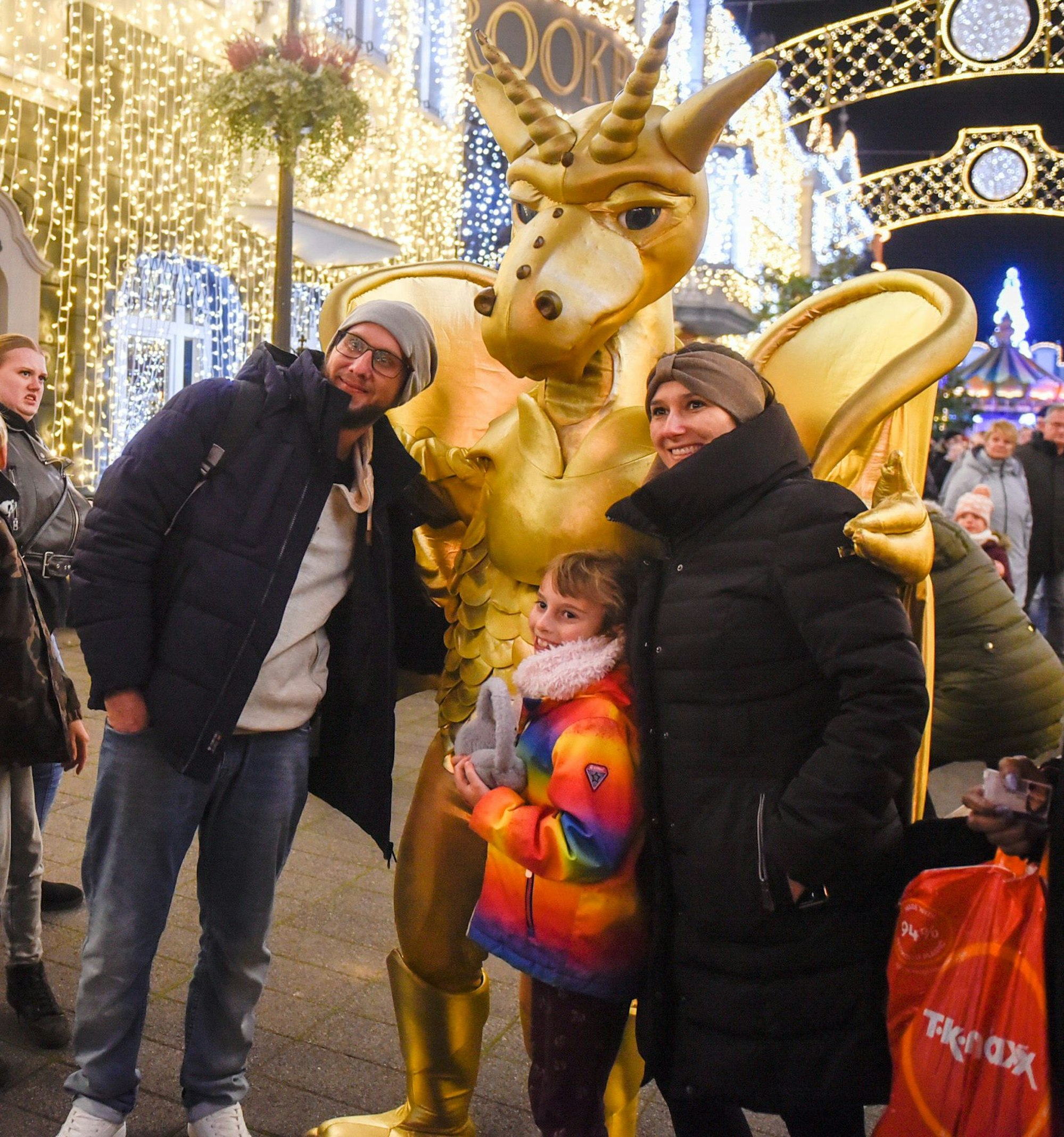 Auf dem Bild ist ein Mädchen zu sehen, das mit ihrer Patentante und deren Mann mit einer goldenen Maskottchen-Figur für ein Foto posiert.