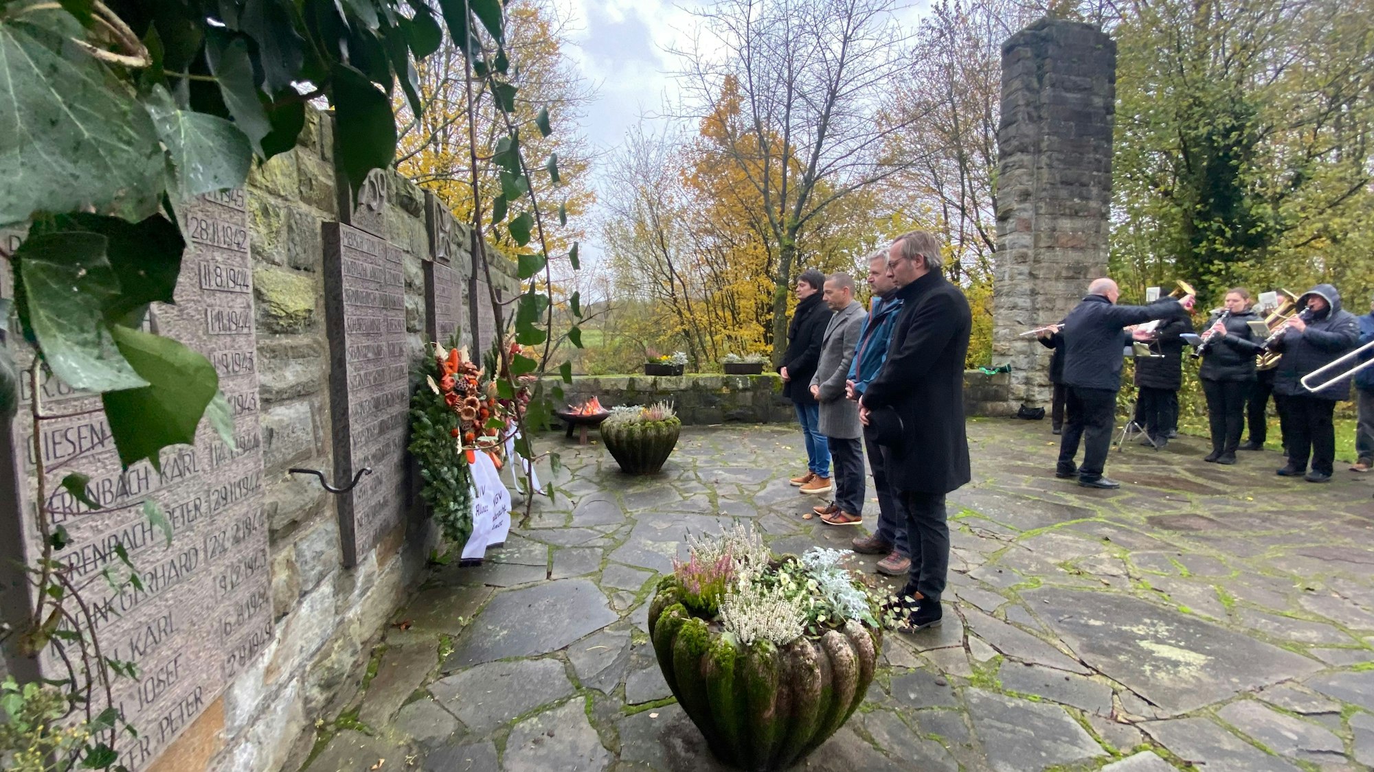 Ein Gefallenendenkmal mit Steinplatten, in denen Namen eingraviert sind. Vier Männer stehen mit verschränkten Händen vor einem niedergelegten Kranz mit Schleifen. Im Hintergrund spielt eine Blaskapelle.
