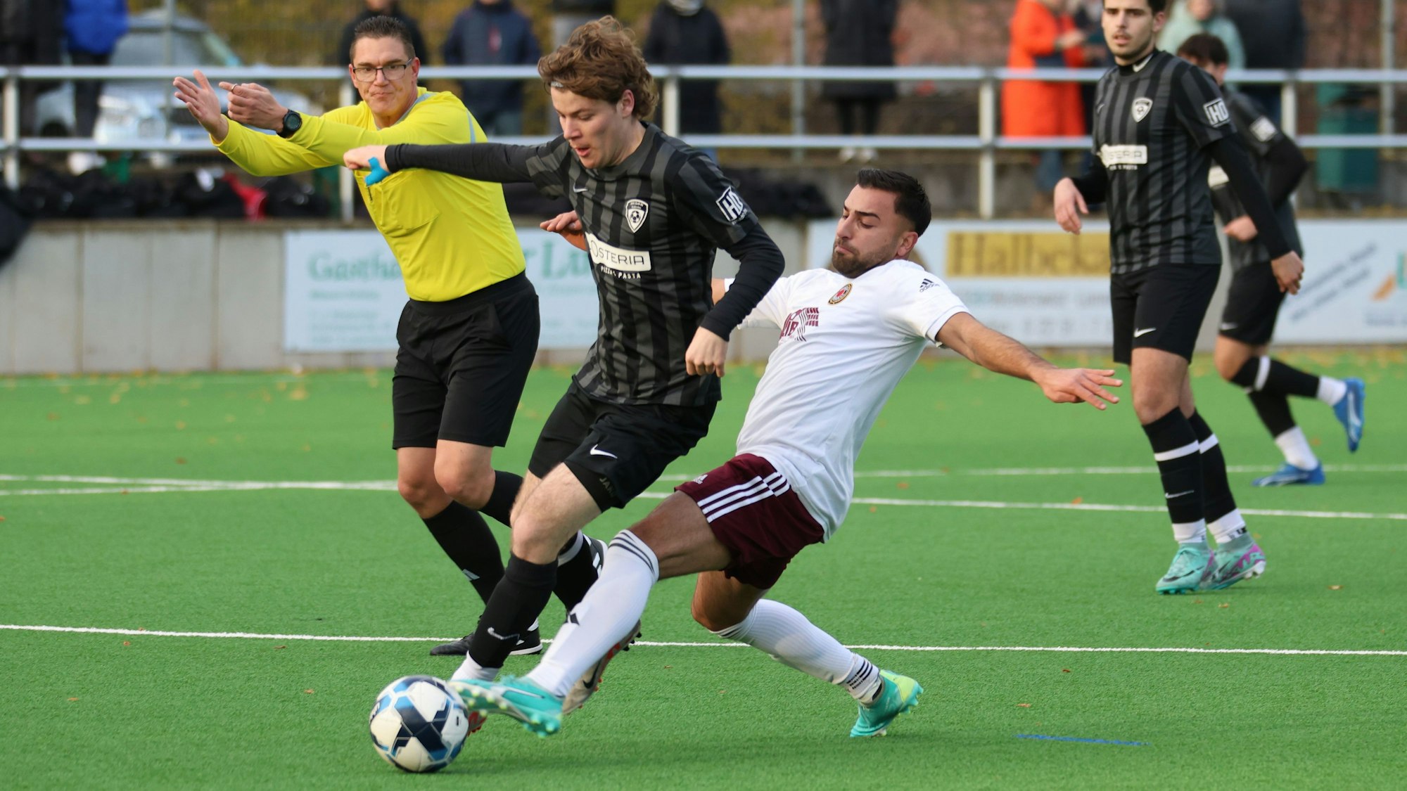 Ein Fußballer grätscht nach einem Ball, der vom Gegenspieler geführt wird. Der Schiedsrichter im Hintergrund deutet Weiterspielen an.