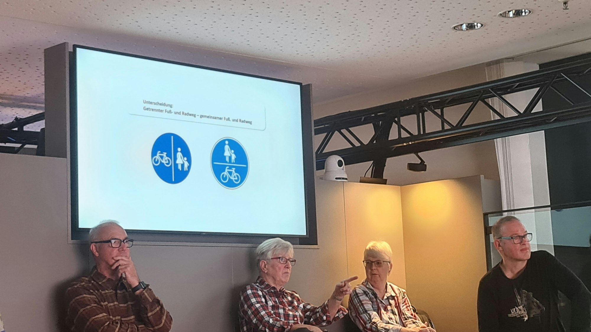 Zu sehen sind vier Teilnehmer des Workshops, die sitzen. Über ihnen ist ein Bildschirm mit zwei unterschiedlichen Verkehrszeichen zu sehen.