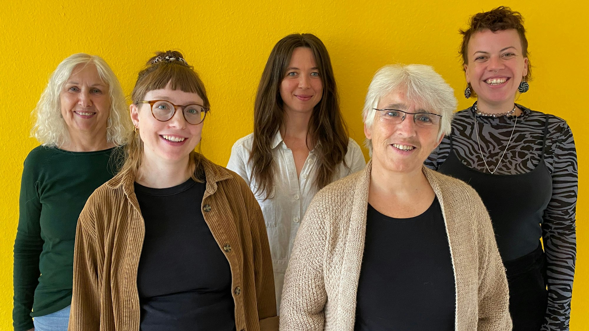 Fünf Frauen unterschiedlichen Alters stehen vor einer gelben Wand und lächeln in die Kamera.