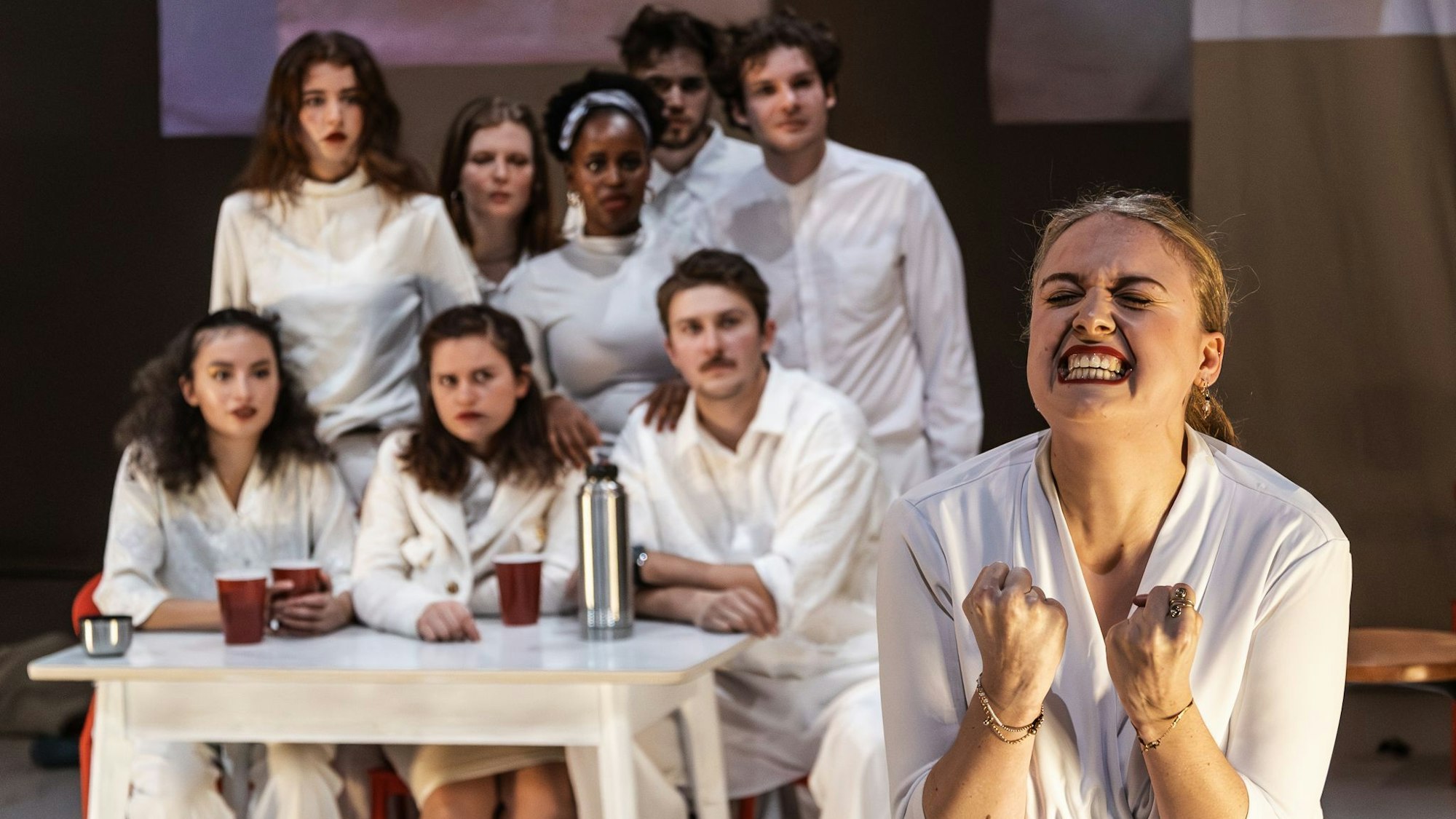 Acht junge Schauspieler und Schauspielerinnen sitzen an und stehen hinter einem Tisch, vorne rechts steht eine Schauspielerin, die die Hände zu Fäusten geballt und das Gesicht verzerrt hat. Alle tragen weiße Kleidung.