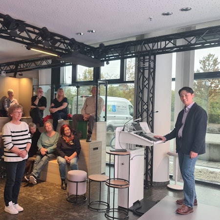 Zu sehen ist Dr. Kim, der auf einer Bühne an einem Rednerpult steht, sowie mehrere Teilnehmerinnen und Teilnehmer des Workshops.