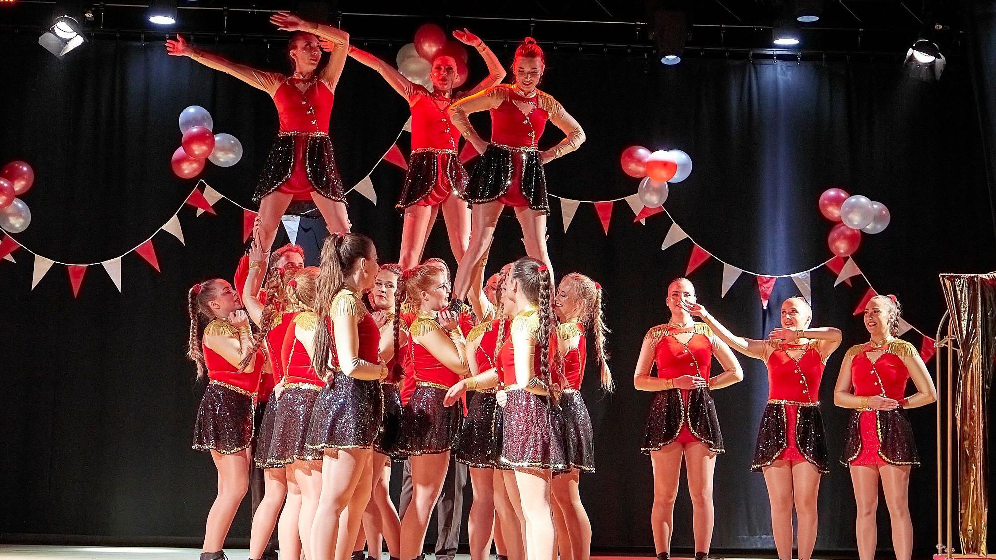 Drei junge Frauen stehen auf den Schultern anderer junger Frauen in Tanzoutfits auf einer Bühne.