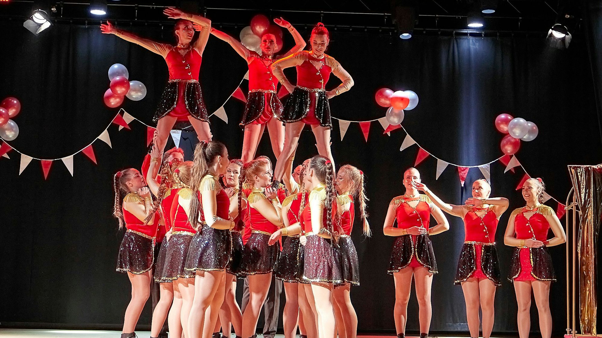 Drei junge Frauen stehen auf den Schultern anderer junger Frauen in Tanzoutfits auf einer Bühne.