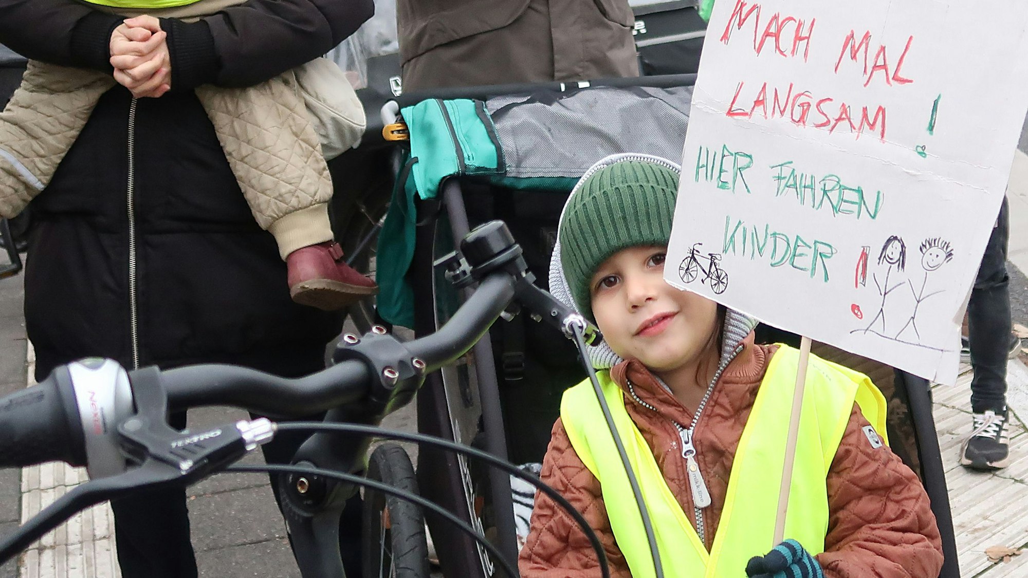 Kind mit Mütze und Warnweste hält Schild mit Aufschrift „Mach mal langsam! Hier fahren Kinder!“ hoch.