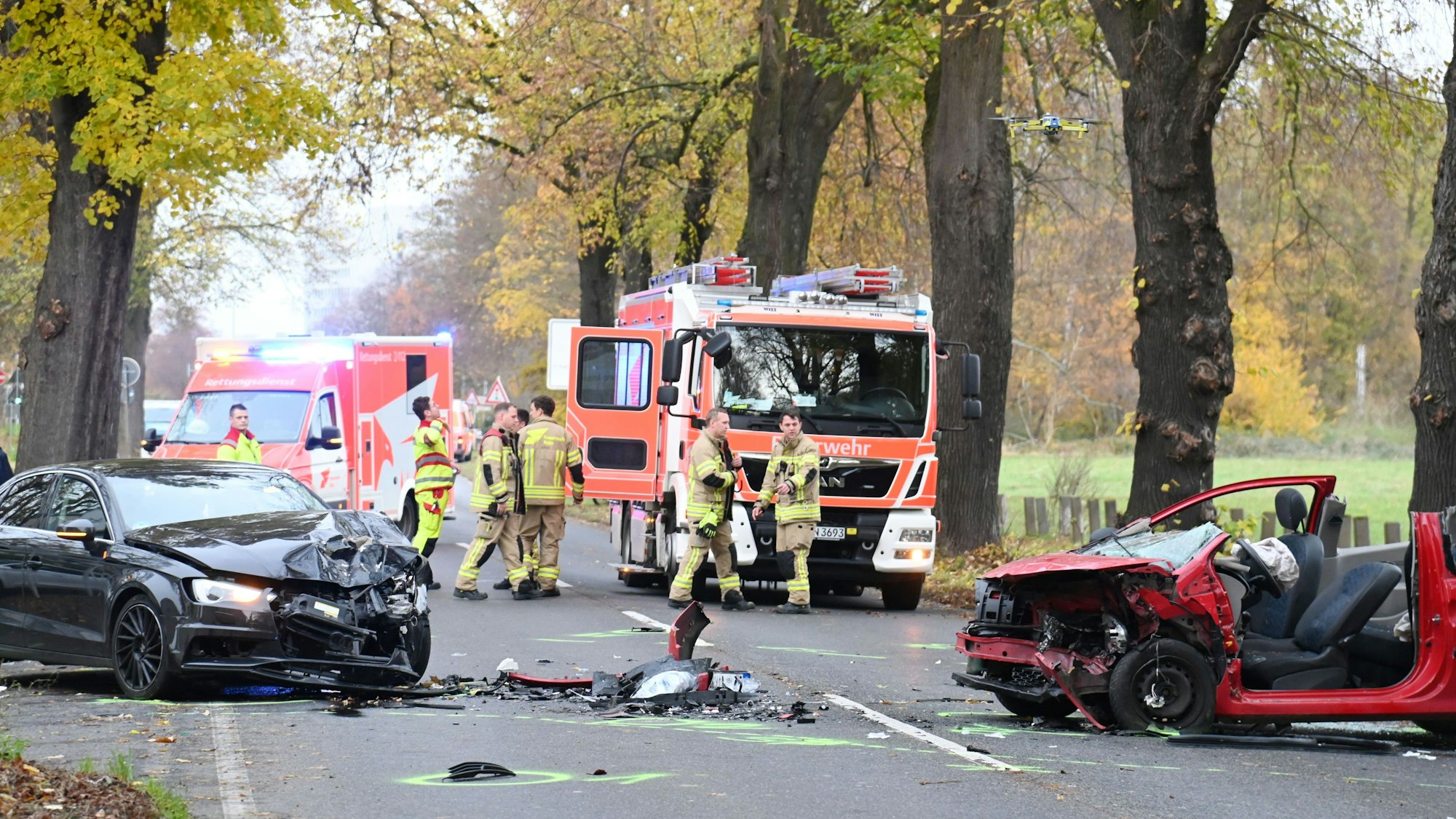 Zwei demolierte Autos stehen auf einer Straße. Bei einem Auto fehlt das komplette Dach. Im Hintergrund stehen ein Feuerwehrwagen und ein Rettungswagen. Einsatzkräfte stehen auf der Straße.